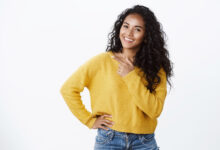 carefree modern young african american girl with curly hair in yellow sweater tilt head joyfully smiling look camera as discuss cool new promo pointing left blank space over white wall