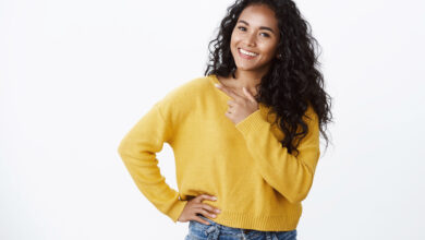 carefree modern young african american girl with curly hair in yellow sweater tilt head joyfully smiling look camera as discuss cool new promo pointing left blank space over white wall