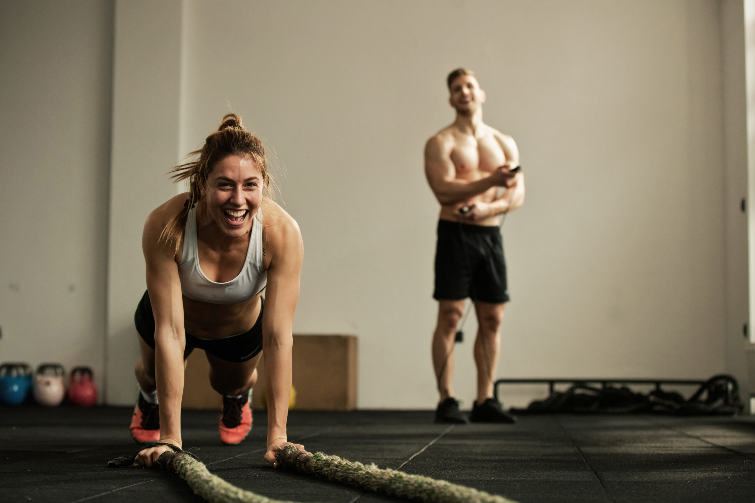 cheerful female athlete doing pushups while exercising with battle rope and having fun in a gym there is a man in the background scaled