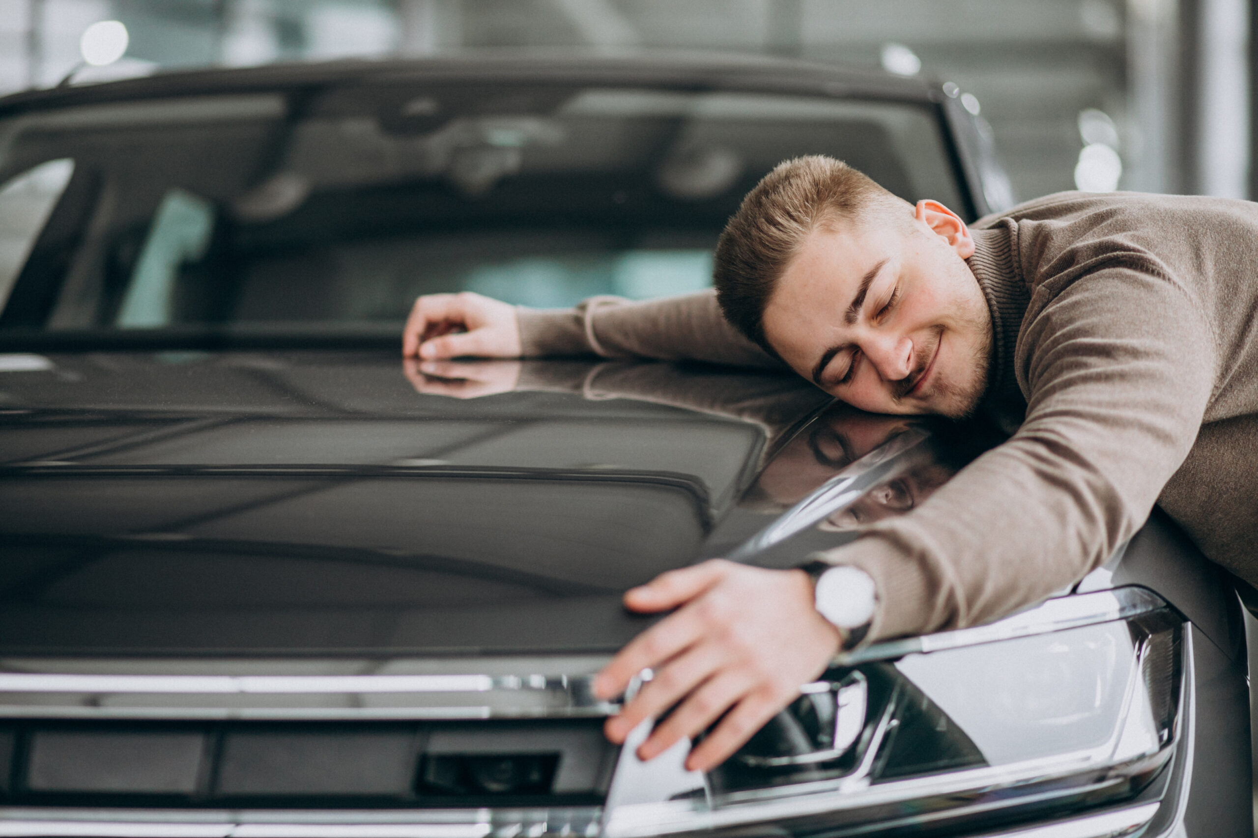 young handsome man hugging a car in a car showroom scaled